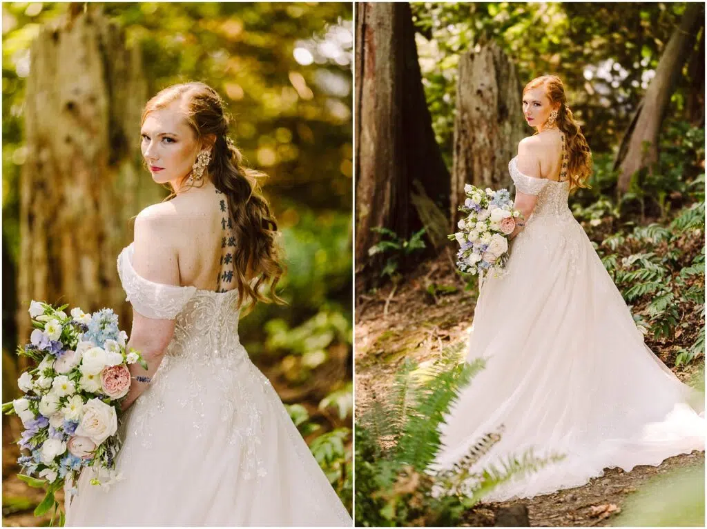Snohomish Wedding Photographer GSquared Weddings A bride with long, wavy red hair stands in a forest, wearing an off-the-shoulder lace wedding gown with a long train. She holds a bouquet of blue, white, and pink flowers and gazes back at the camera. Sunlight filters through the trees. Serving Seattle, Snohomish and Orlando