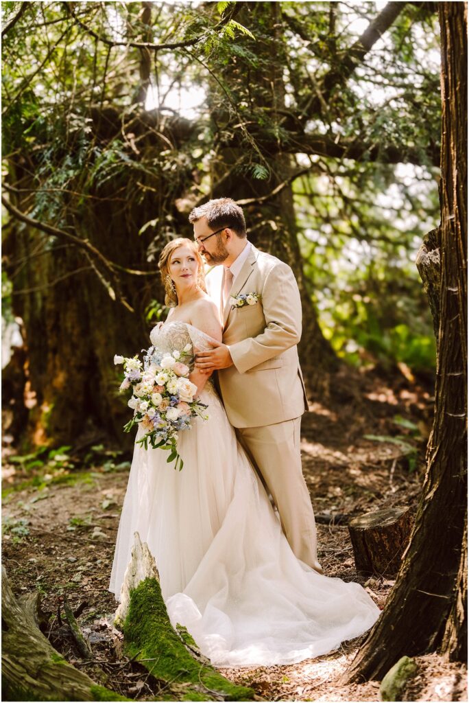Snohomish Wedding Photographer GSquared Weddings Bride and groom stand together in a wooded area. The bride, holding a bouquet of flowers, wears a white gown, while the groom is in a light tan suit. Sunlight filters through the trees, creating a serene and romantic atmosphere. Serving Seattle, Snohomish and Orlando