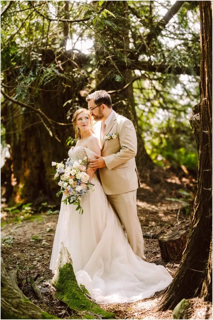 Snohomish Wedding Photographer GSquared Weddings Bride and groom stand together in a wooded area. The bride, holding a bouquet of flowers, wears a white gown, while the groom is in a light tan suit. Sunlight filters through the trees, creating a serene and romantic atmosphere. Serving Seattle, Snohomish and Orlando