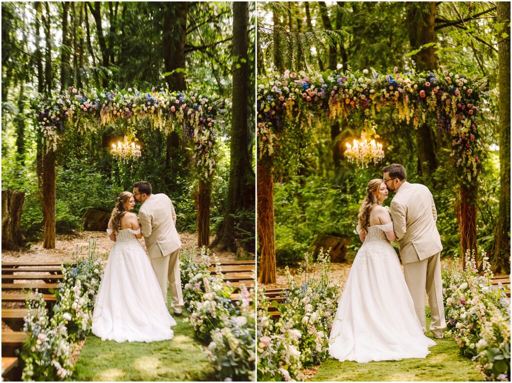 Snohomish Wedding Photographer GSquared Weddings A couple shares a kiss during their wedding ceremony in a lush, wooded setting. They stand under a floral arch adorned with greenery and flowers. The aisle is lined with floral arrangements, and a chandelier hangs above them. Serving Seattle, Snohomish and Orlando