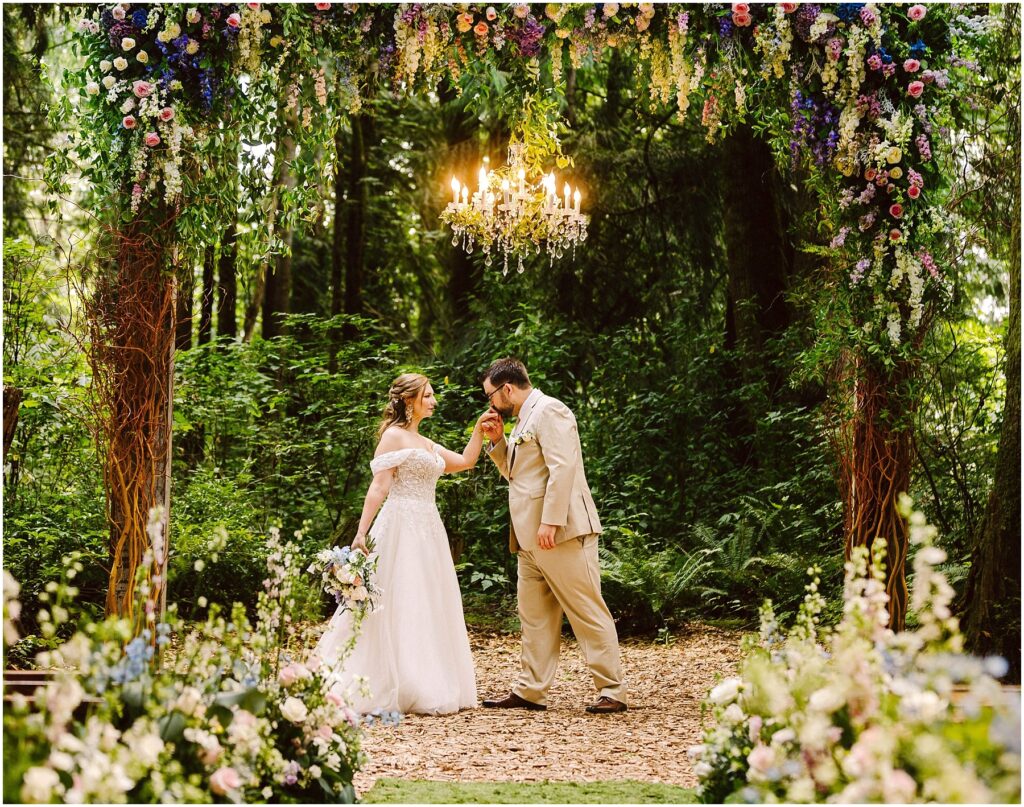 Snohomish Wedding Photographer GSquared Weddings A bride in a white dress and groom in a beige suit stand under a floral arch in a forest. The groom kisses the brides hand. A chandelier hangs above them, and colorful flowers surround the scene. Serving Seattle, Snohomish and Orlando