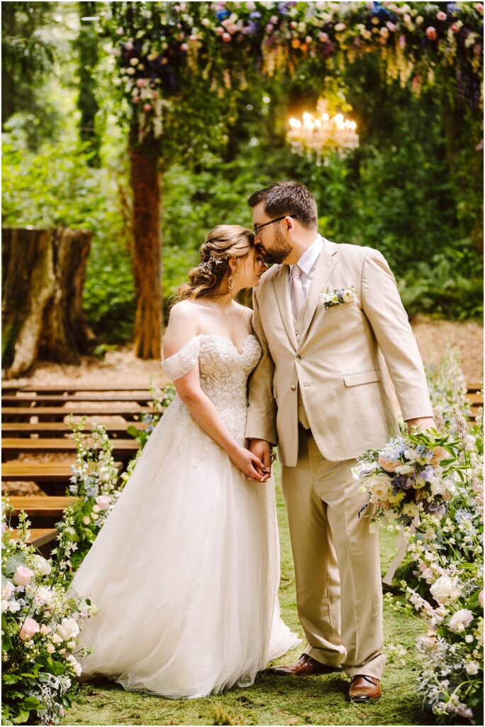 Snohomish Wedding Photographer GSquared Weddings A bride and groom hold hands and lean in for a kiss in an outdoor wedding setting. The bride wears an off-shoulder gown, and the groom dons a beige suit. They are surrounded by greenery and floral arrangements, with a chandelier hanging above. Serving Seattle, Snohomish and Orlando