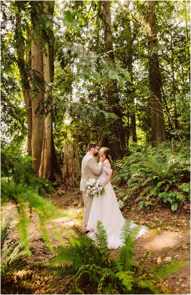 Snohomish Wedding Photographer GSquared Weddings A bride and groom share a kiss in a lush forest setting. The bride holds a bouquet, and sunlight filters through the tall trees and leafy greenery, creating a serene and romantic atmosphere. Serving Seattle, Snohomish and Orlando