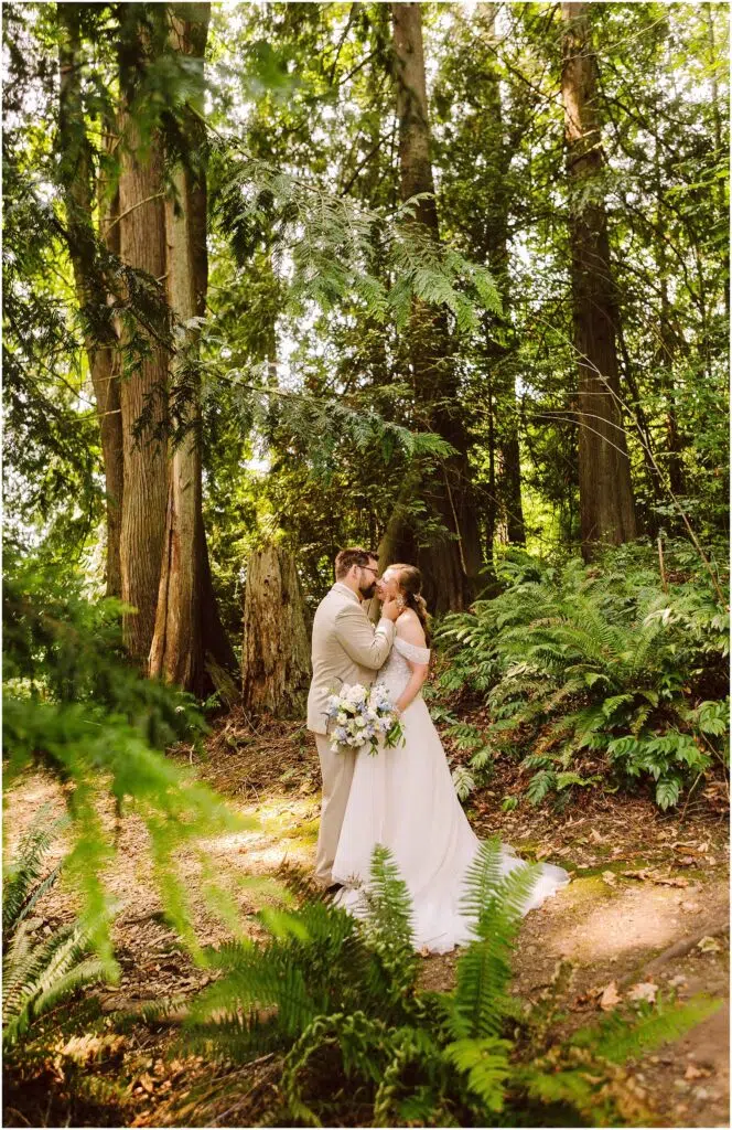 Snohomish Wedding Photographer GSquared Weddings A bride and groom share a kiss in a lush forest setting. The bride holds a bouquet, and sunlight filters through the tall trees and leafy greenery, creating a serene and romantic atmosphere. Serving Seattle, Snohomish and Orlando
