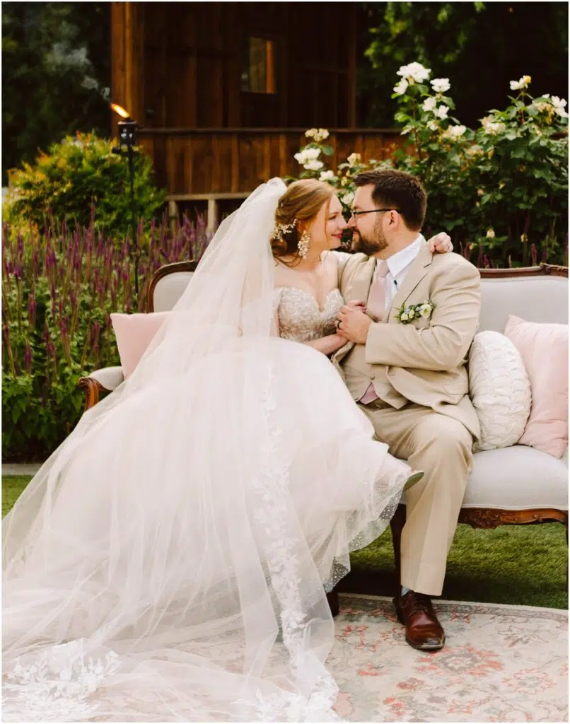 Snohomish Wedding Photographer GSquared Weddings A bride and groom sit closely on an outdoor sofa surrounded by flowers. The bride wears a white dress with a long veil, and the groom wears a tan suit. They gaze into each others eyes, holding hands, with a wooden building in the background. Serving Seattle, Snohomish and Orlando