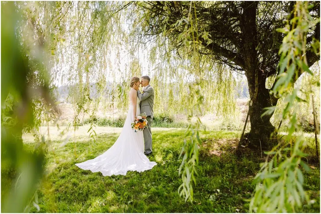 Snohomish Wedding Photographer GSquared Weddings A bride and groom share a tender moment under a large, leafy tree. The bride wears a long white gown and holds a bouquet of colorful flowers. Sunlight filters through the branches, casting dappled shadows on the grassy ground. Serving Seattle, Snohomish and Orlando