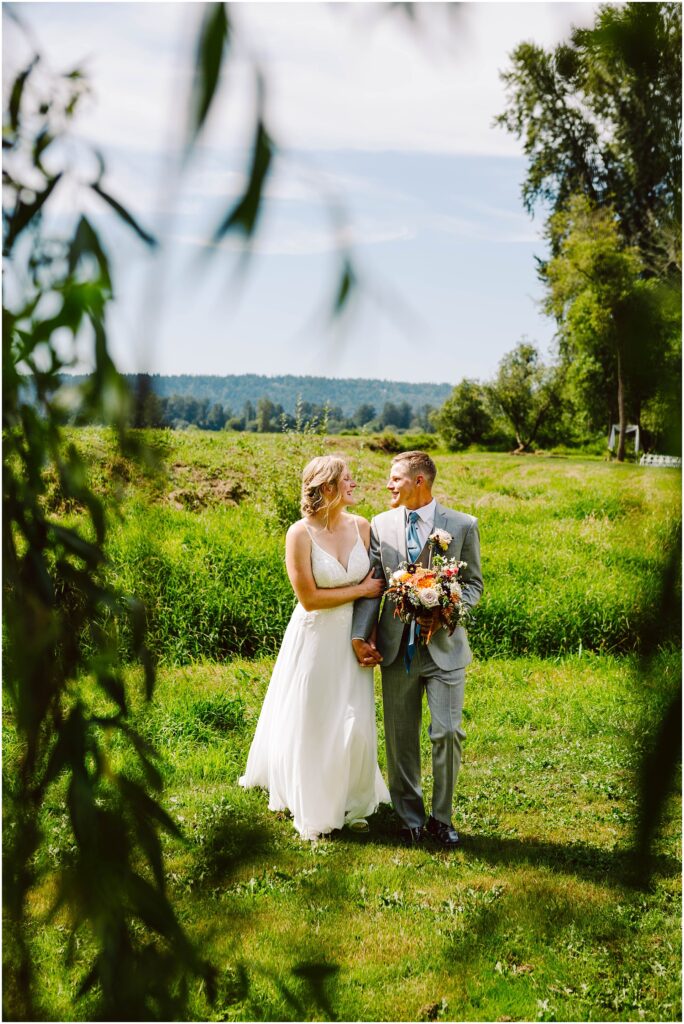 Snohomish Wedding Photographer GSquared Weddings A couple stands in a lush, green outdoor setting. The woman wears a white dress and the man a gray suit. They hold hands and smile at each other, with flowers in their hands. Trees and a clear blue sky are in the background. Serving Seattle, Snohomish and Orlando