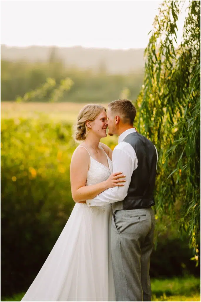 Snohomish Wedding Photographer GSquared Weddings A couple stands outdoors in a lush garden setting during sunset. The woman wears a white wedding dress and the man is in a shirt, vest, and pants. They embrace and smile at each other, surrounded by greenery and soft, warm light. Serving Seattle, Snohomish and Orlando