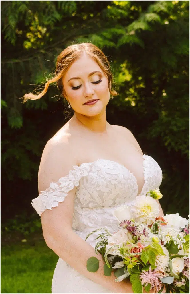 Snohomish Wedding Photographer GSquared Weddings Bride in an off-shoulder lace wedding dress holds a bouquet of mixed flowers. She stands outdoors against a backdrop of lush greenery, with her eyes closed and a serene expression, as a wisp of hair gently falls across her face. Serving Seattle, Snohomish and Orlando