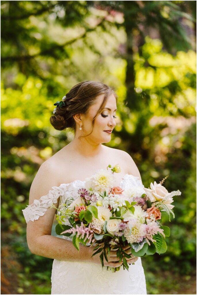 Snohomish Wedding Photographer GSquared Weddings A bride in a lace off-shoulder wedding dress stands outdoors holding a colorful bouquet. Her hair is styled in an updo, adorned with greenery. The background is a lush, sunlit garden with green foliage. Serving Seattle, Snohomish and Orlando