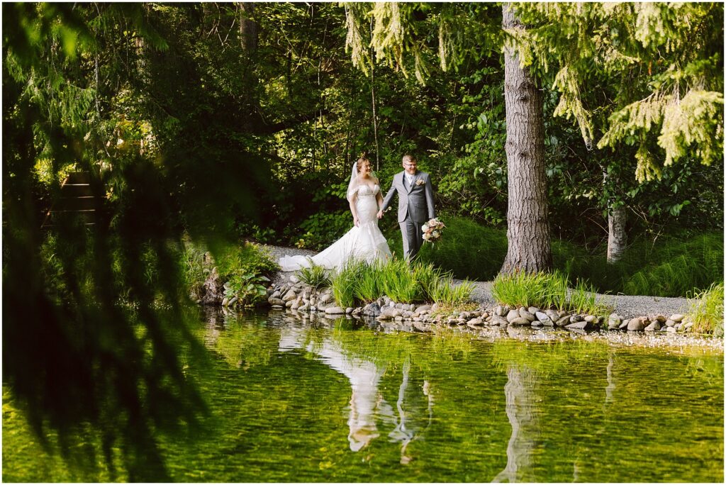 Snohomish Wedding Photographer GSquared Weddings A bride in a white gown and a groom in a gray suit walk hand in hand on a path beside a serene green pond surrounded by lush trees and vegetation. Their reflection is visible in the water. Serving Seattle, Snohomish and Orlando