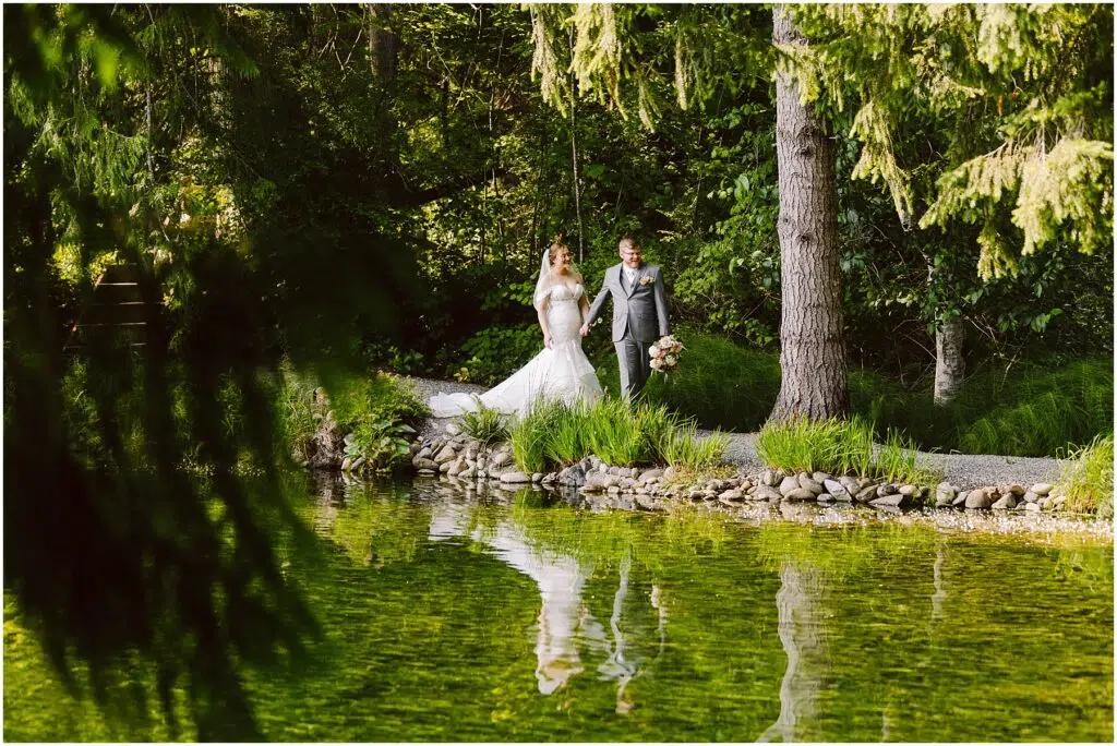Snohomish Wedding Photographer GSquared Weddings A bride in a white gown and a groom in a gray suit walk hand in hand on a path beside a serene green pond surrounded by lush trees and vegetation. Their reflection is visible in the water. Serving Seattle, Snohomish and Orlando