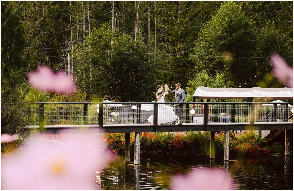 Snohomish Wedding Photographer GSquared Weddings A bride and groom walk across a wooden bridge over a pond, surrounded by lush greenery and colorful flowers. The background features tall trees, and soft pink flowers are in the foreground, partially framing the scene. Serving Seattle, Snohomish and Orlando