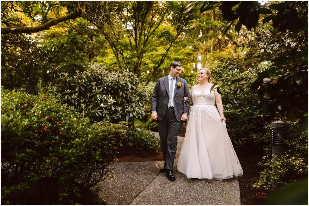 Snohomish Wedding Photographer GSquared Weddings A bride and groom walk arm in arm down a garden path. The bride is wearing a white dress and the groom is in a suit with a yellow boutonniere. They are surrounded by lush green foliage and appear to be smiling. Serving Seattle, Snohomish and Orlando