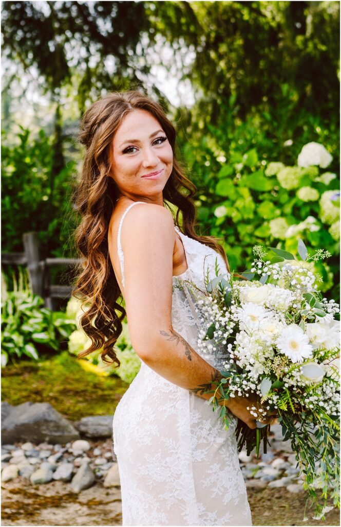 Snohomish Wedding Photographer GSquared Weddings A bride in a white lace wedding dress stands outdoors, holding a bouquet of white flowers and greenery. She has long, wavy brown hair and is smiling slightly. The background features lush greenery and a wooden fence. Serving Seattle, Snohomish and Orlando