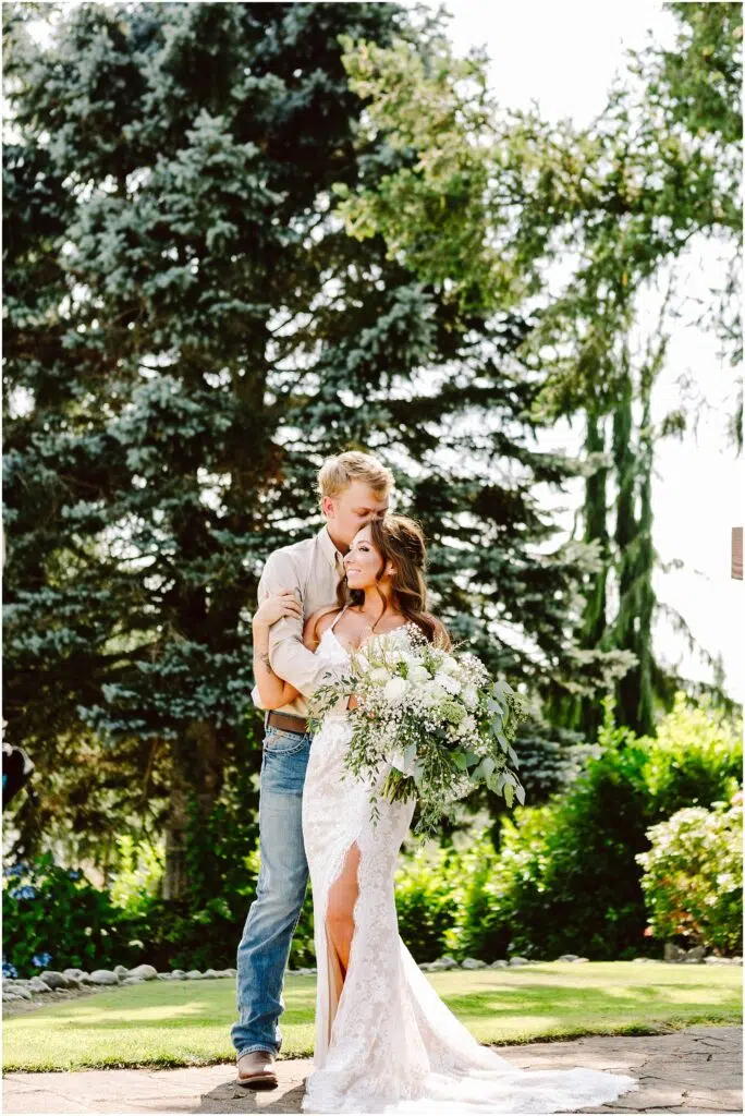 Snohomish Wedding Photographer GSquared Weddings A couple embraces outdoors in front of lush greenery and large trees. The woman, in a white lace wedding dress holding a bouquet, stands closely with a man in a beige shirt and jeans. Sunlight filters through the foliage, creating a serene ambiance. Serving Seattle, Snohomish and Orlando