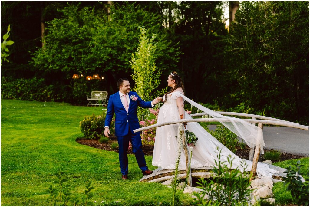 Snohomish Wedding Photographer GSquared Weddings A bride in a white gown and long veil stands on a small wooden bridge, holding hands with a groom in a blue suit and floral tie. They are in a lush green garden with blooming flowers and trees. Serving Seattle, Snohomish and Orlando
