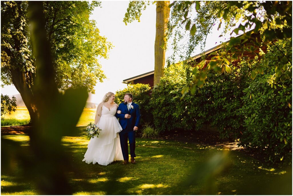 Snohomish Wedding Photographer GSquared Weddings A bride and groom walk hand in hand through a sunlit garden surrounded by lush green trees and bushes. The bride wears a white dress, and the groom is in a dark suit. Sunlight filters through the leaves, casting gentle shadows. Serving Seattle, Snohomish and Orlando