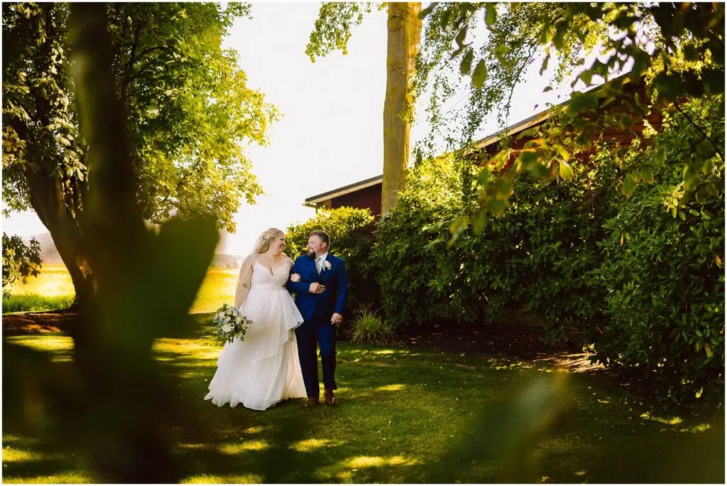 Snohomish Wedding Photographer GSquared Weddings A bride and groom walk hand in hand through a sunlit garden surrounded by lush green trees and bushes. The bride wears a white dress, and the groom is in a dark suit. Sunlight filters through the leaves, casting gentle shadows. Serving Seattle, Snohomish and Orlando