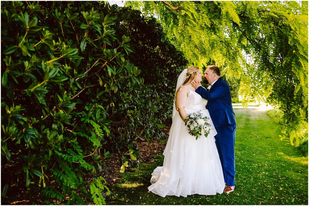 Snohomish Wedding Photographer GSquared Weddings A bride in a white gown and veil, holding a bouquet of blue and white flowers, gazes lovingly at a groom in a blue suit. They stand close under lush green trees with sunlight filtering through the leaves, creating a romantic, serene setting. Serving Seattle, Snohomish and Orlando