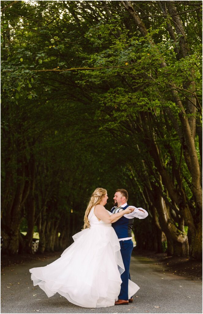 Snohomish Wedding Photographer GSquared Weddings A couple dances on a road under a canopy of lush green trees. The woman wears a flowing white gown, and the man is in a blue suit. They are surrounded by a serene, verdant environment, creating a romantic and whimsical atmosphere. Serving Seattle, Snohomish and Orlando