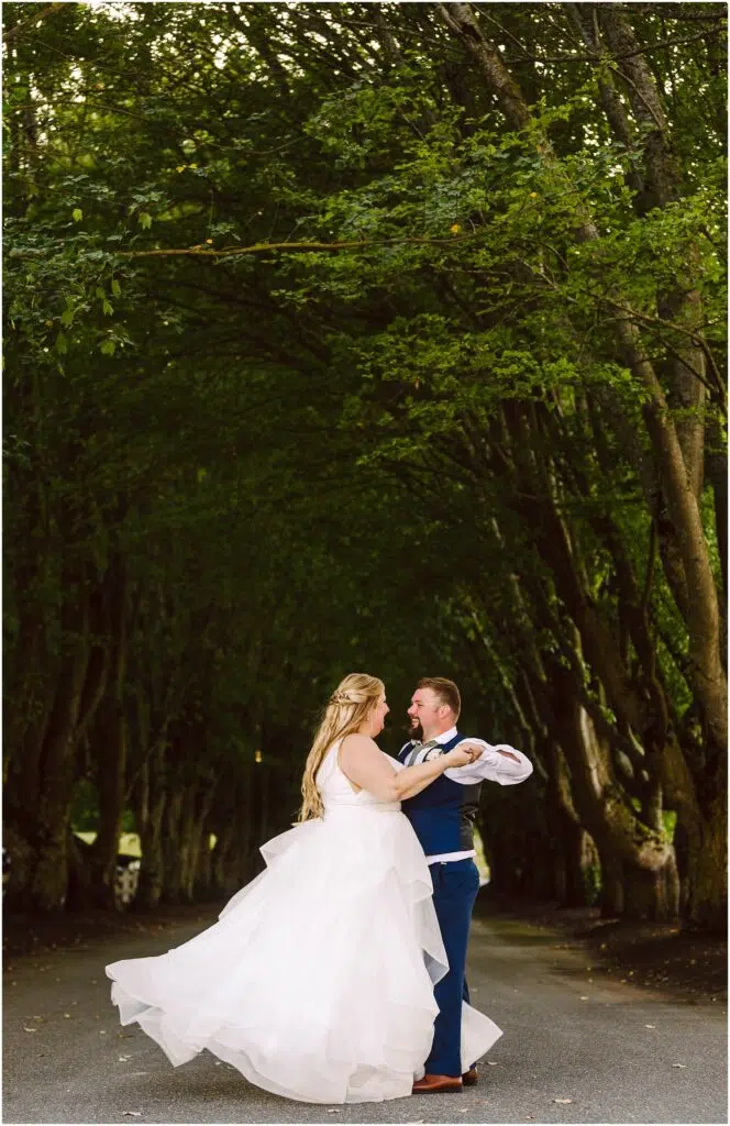Snohomish Wedding Photographer GSquared Weddings A couple dances on a road under a canopy of lush green trees. The woman wears a flowing white gown, and the man is in a blue suit. They are surrounded by a serene, verdant environment, creating a romantic and whimsical atmosphere. Serving Seattle, Snohomish and Orlando