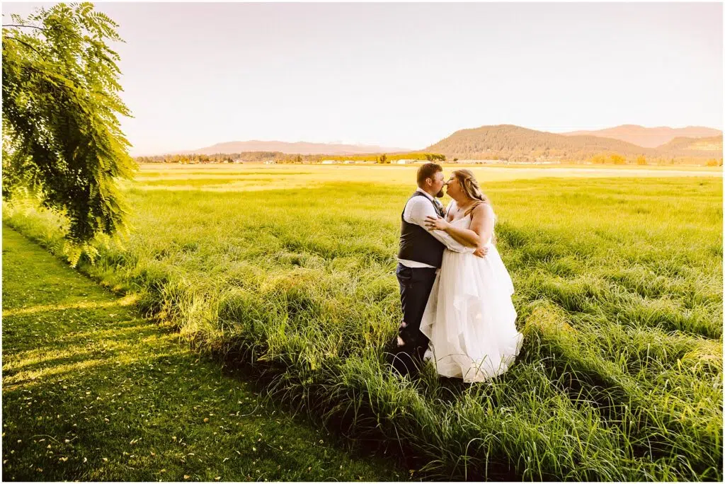 Snohomish Wedding Photographer GSquared Weddings A bride and groom stand embracing in a green field under a clear sky. The bride wears a white dress, and the groom is in a vest and tie. A tree and distant mountains frame the idyllic scene. Serving Seattle, Snohomish and Orlando