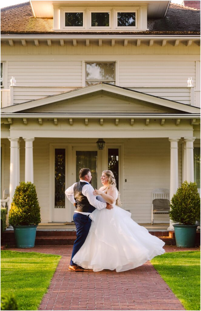 Snohomish Wedding Photographer GSquared Weddings A couple dances joyfully on a red-brick path in front of a large, white house with columns. The bride wears a flowing white gown, and the groom is in a vest and pants. Both are smiling amidst a backdrop of green grass and potted shrubs. Serving Seattle, Snohomish and Orlando