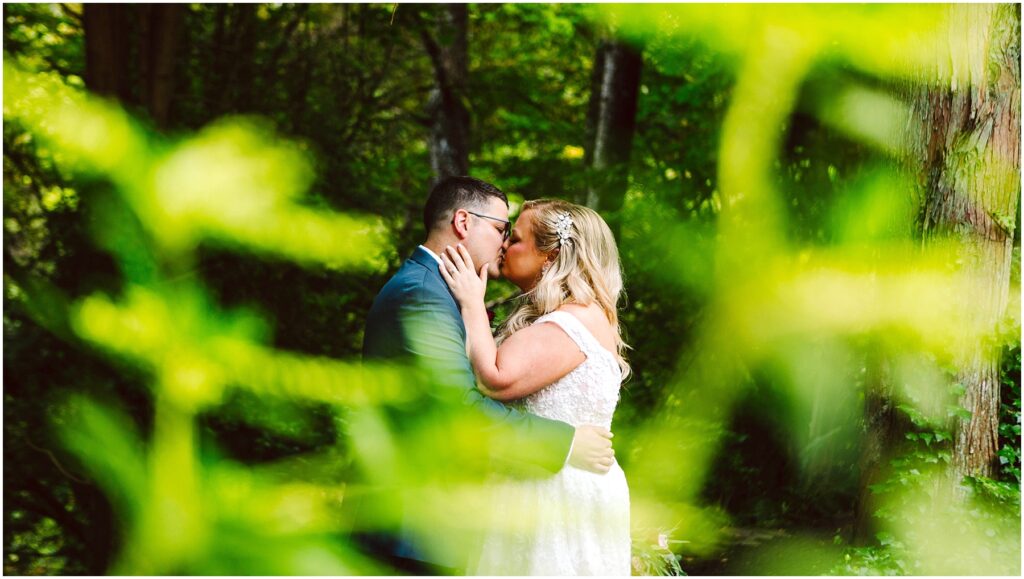 Snohomish Wedding Photographer GSquared Weddings A couple is kissing in a lush, green forest. The bride is wearing a white dress, and the groom is in a suit. The scene is partially framed by out-of-focus greenery, creating a vibrant and intimate atmosphere. Serving Seattle, Snohomish and Orlando
