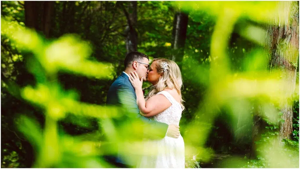 Snohomish Wedding Photographer GSquared Weddings A couple is kissing in a lush, green forest. The bride is wearing a white dress, and the groom is in a suit. The scene is partially framed by out-of-focus greenery, creating a vibrant and intimate atmosphere. Serving Seattle, Snohomish and Orlando