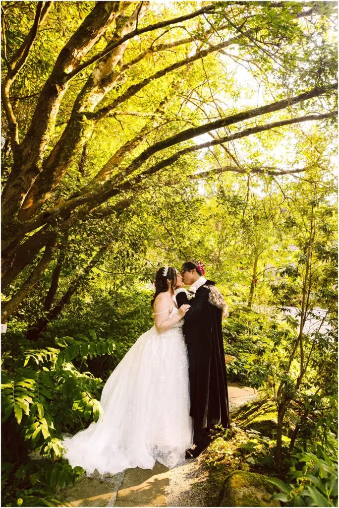 Snohomish Wedding Photographer GSquared Weddings A couple in wedding attire stand together in a lush, sunlit garden. The bride wears a white dress and the groom a black suit. They appear to be sharing an intimate moment surrounded by vibrant greenery and dappled sunlight filtering through the trees. Serving Seattle, Snohomish and Orlando