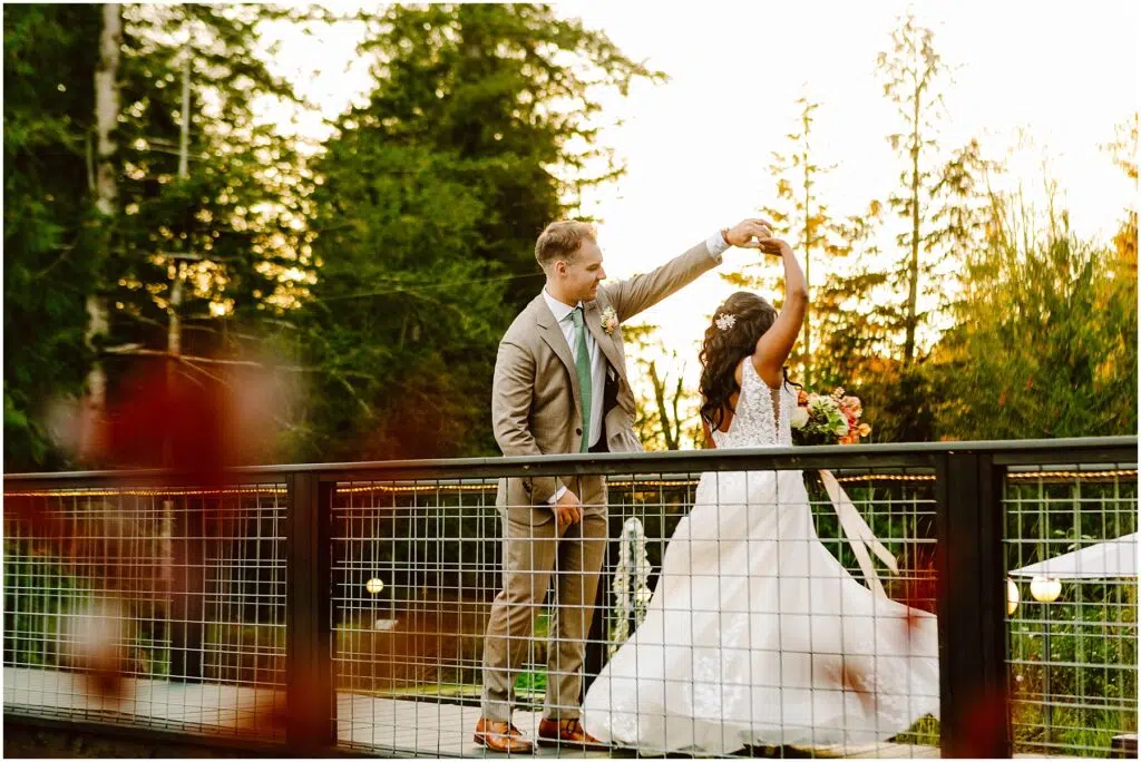 Snohomish Wedding Photographer GSquared Weddings A groom in a brown suit twirls his bride, who is wearing a white wedding dress, on a wooden bridge. They are surrounded by trees, with a warm sunset glow in the background. Serving Seattle, Snohomish and Orlando