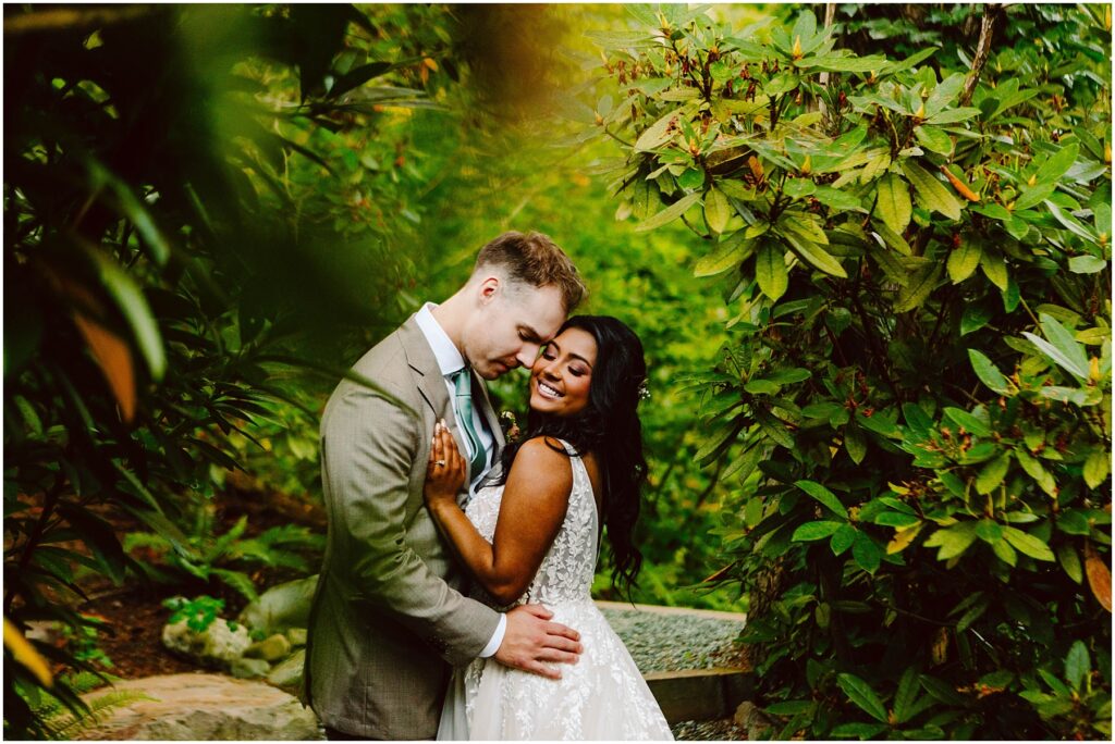 Snohomish Wedding Photographer GSquared Weddings A couple embraces in a lush, green garden. The man in a gray suit gently leans his forehead against the woman, who smiles while wearing a white lace dress. They are surrounded by vibrant foliage, creating a serene, romantic atmosphere. Serving Seattle, Snohomish and Orlando