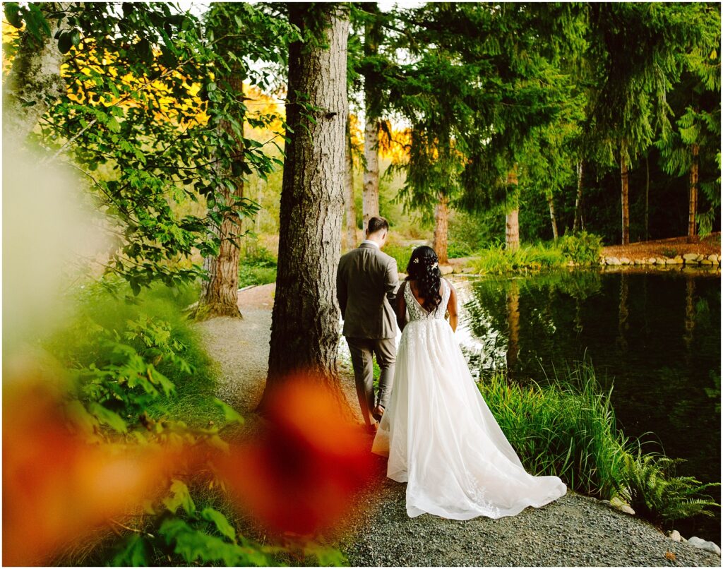 Snohomish Wedding Photographer GSquared Weddings Bride and groom walking along a wooded path beside a serene lake, surrounded by lush greenery and tall trees. The bride is in a white dress, and the groom is in a suit. The scene is vibrant with hints of autumn colors. Serving Seattle, Snohomish and Orlando