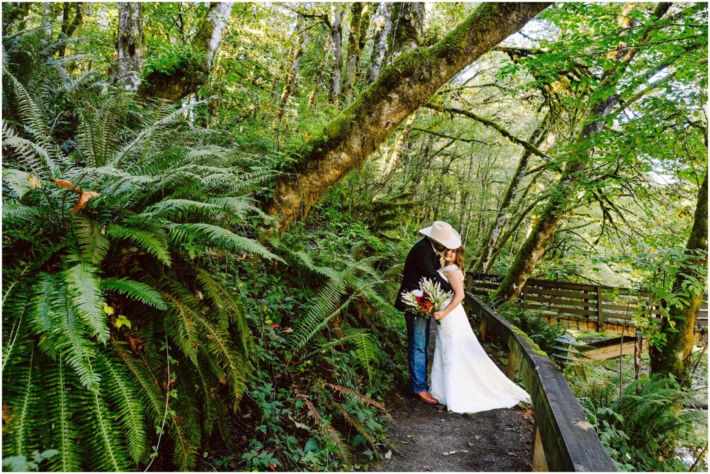 Snohomish Wedding Photographer GSquared Weddings A bride in a white dress and a groom in a cowboy hat embrace on a forest path surrounded by lush green ferns and tall trees. A wooden bridge is visible in the background under dappled sunlight. Serving Seattle, Snohomish and Orlando
