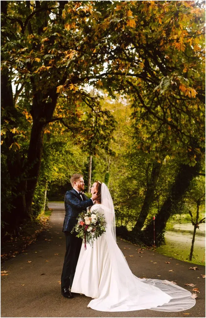 Snohomish Wedding Photographer GSquared Weddings A couple in wedding attire embrace on a tree-lined path. The bride wears a white gown and veil, holding a bouquet, while the groom is in a dark suit. Autumn leaves add color to the scene, with green and golden foliage overhead. Serving Seattle, Snohomish and Orlando