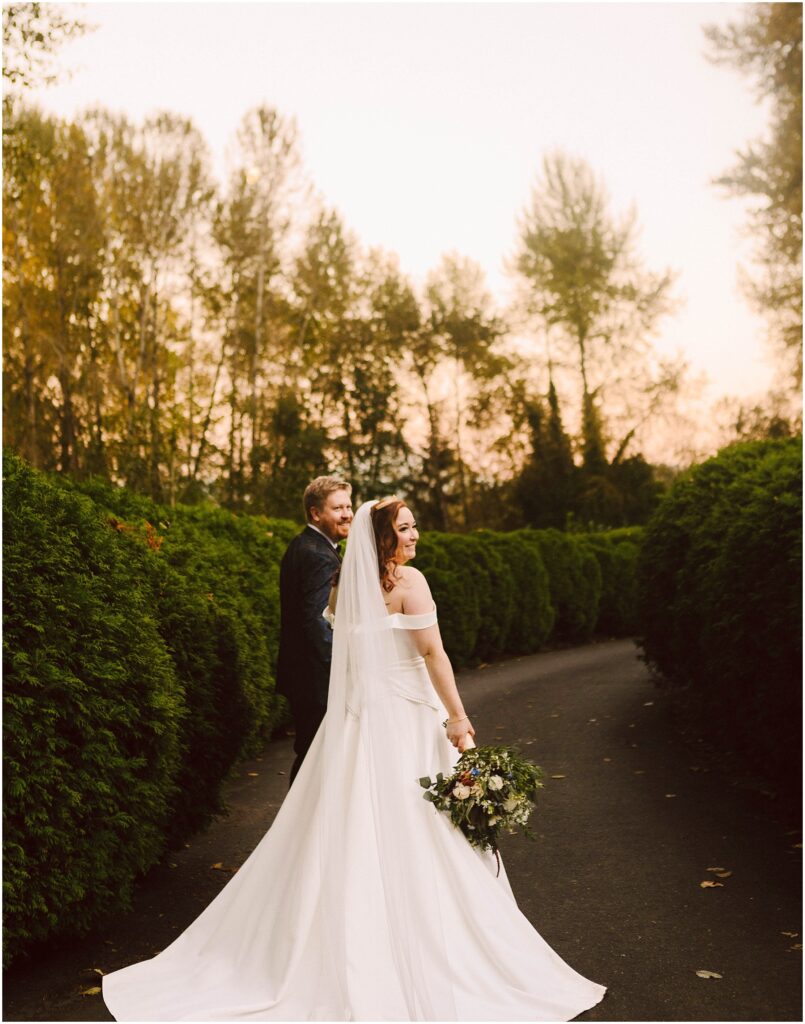 Snohomish Wedding Photographer GSquared Weddings Bride in an off-shoulder gown and veil holds a bouquet while walking hand-in-hand with a groom in a dark suit. They stroll on a tree-lined path at sunset, looking back and smiling. Serving Seattle, Snohomish and Orlando