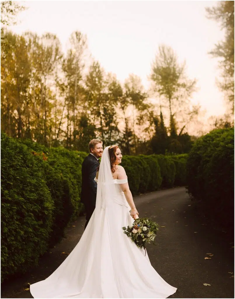 Snohomish Wedding Photographer GSquared Weddings Bride in an off-shoulder gown and veil holds a bouquet while walking hand-in-hand with a groom in a dark suit. They stroll on a tree-lined path at sunset, looking back and smiling. Serving Seattle, Snohomish and Orlando