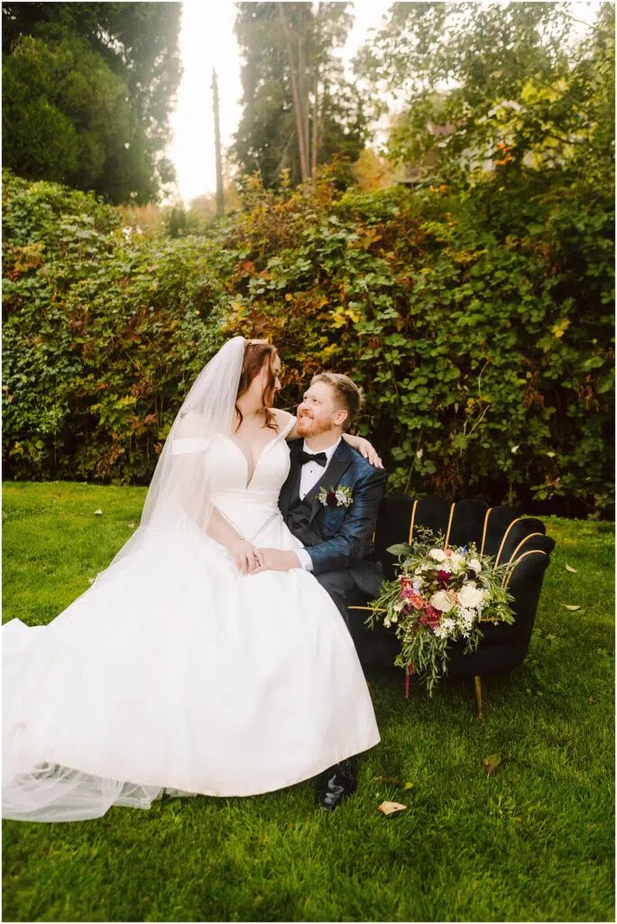 Snohomish Wedding Photographer GSquared Weddings A bride in a white dress and veil sits beside a groom in a black suit and bowtie on a chair outdoors. They are smiling at each other. A vibrant bouquet is placed on the chair. There is greenery in the background. Serving Seattle, Snohomish and Orlando