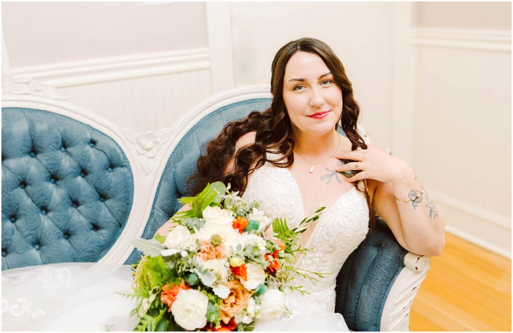 Snohomish Wedding Photographer GSquared Weddings Bride in a white lace wedding dress sits on a vintage blue couch, holding a bouquet of white, orange, and green flowers. She has long brown hair and is smiling softly, with tattoos visible on her arms. Serving Seattle, Snohomish and Orlando