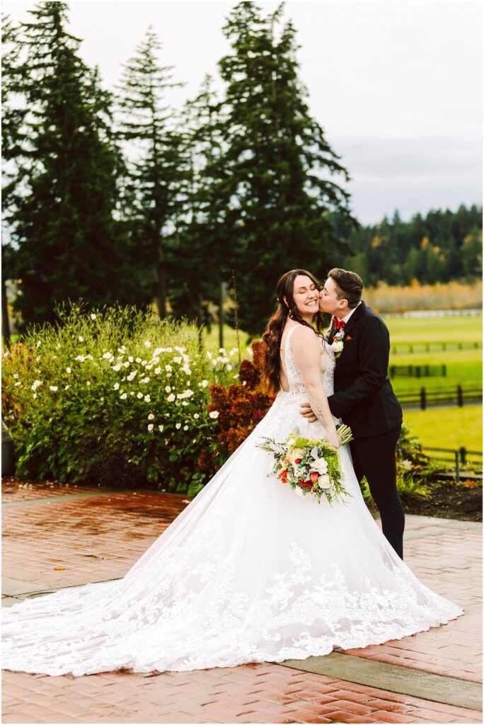 Snohomish Wedding Photographer GSquared Weddings A bride in a white lace gown holds a bouquet while smiling, as a person in a dark suit kisses her cheek. They stand on a wet patio, surrounded by greenery and trees. The scene is outdoors on a cloudy day. Serving Seattle, Snohomish and Orlando