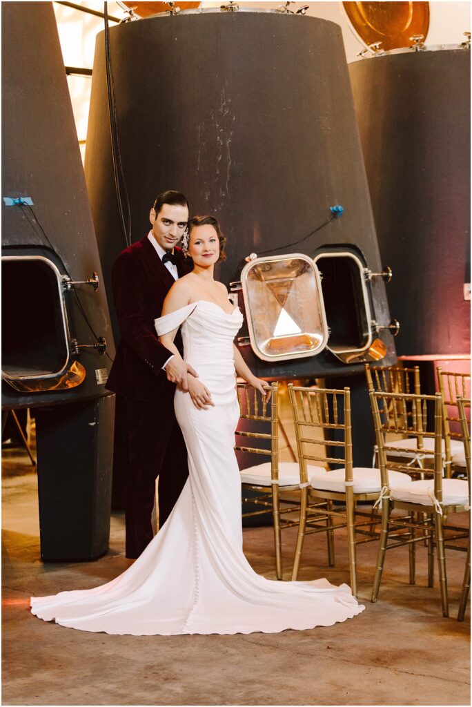 Snohomish Wedding Photographer GSquared Weddings A couple stands in front of large industrial tanks. The woman wears an off-shoulder white gown, and the man wears a burgundy suit. They are surrounded by rows of chairs, suggesting a wedding or formal event setting. Serving Seattle, Snohomish and Orlando