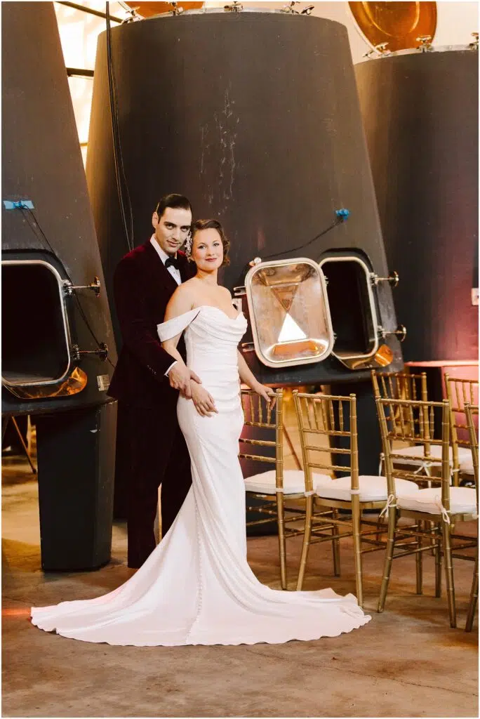 Snohomish Wedding Photographer GSquared Weddings A couple stands in front of large industrial tanks. The woman wears an off-shoulder white gown, and the man wears a burgundy suit. They are surrounded by rows of chairs, suggesting a wedding or formal event setting. Serving Seattle, Snohomish and Orlando