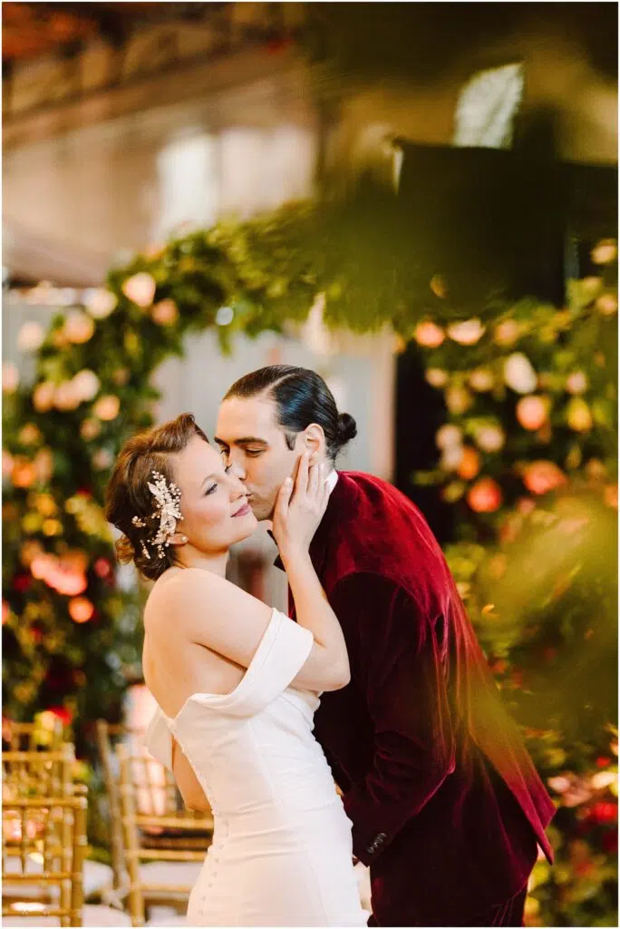 Snohomish Wedding Photographer GSquared Weddings A couple shares a tender moment at a wedding ceremony. The woman wears a white dress with off-the-shoulder sleeves and a decorative hairpiece, while the man wears a red velvet suit. They are surrounded by lush greenery and floral arrangements. Serving Seattle, Snohomish and Orlando