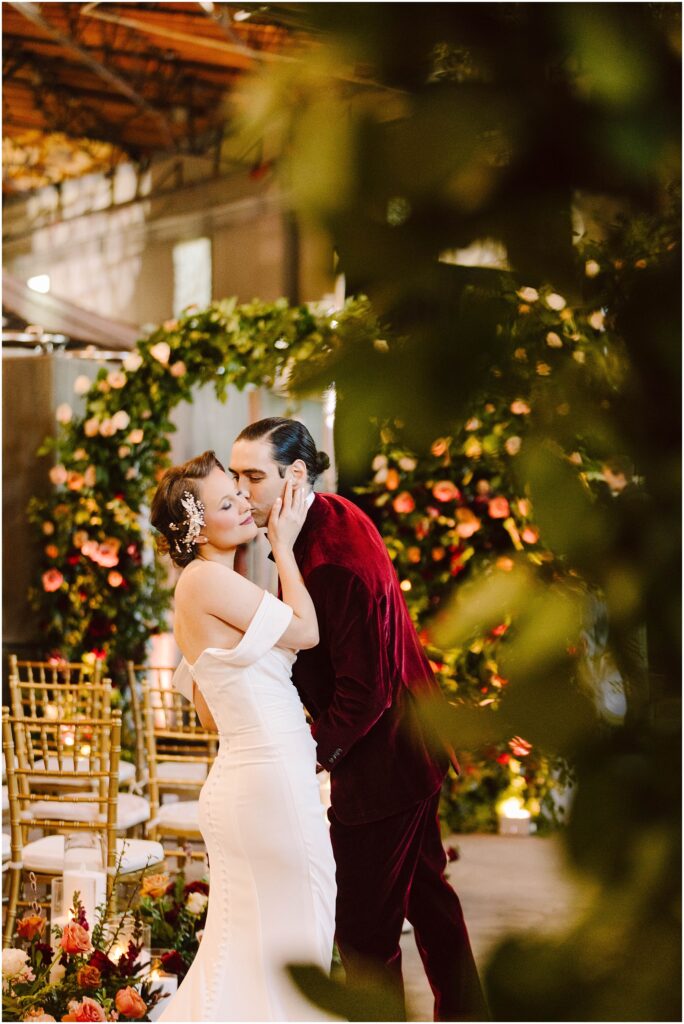 Snohomish Wedding Photographer GSquared Weddings A couple shares an intimate moment during their wedding. The bride is in a white gown with floral hair accessories, while the groom wears a burgundy suit. Theyre surrounded by floral decorations and gold chairs. Serving Seattle, Snohomish and Orlando