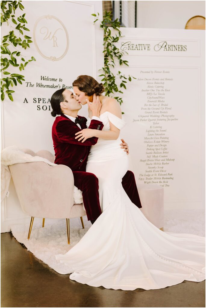 Snohomish Wedding Photographer GSquared Weddings A couple in wedding attire shares a kiss. The person in a white gown is seated on a plush chair, while the person in a burgundy suit kneels beside them. Behind them is a decorated panel with writing and green foliage. Serving Seattle, Snohomish and Orlando