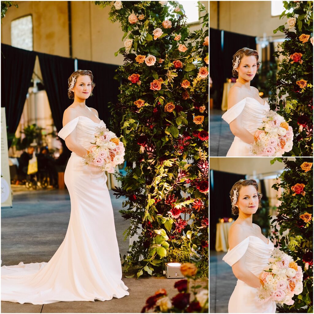 Snohomish Wedding Photographer GSquared Weddings A bride in an off-shoulder white dress holds a bouquet of pastel flowers. She stands beside a tall floral arrangement with roses and greenery. The setting is indoors with dark drapery and warm lighting. Serving Seattle, Snohomish and Orlando