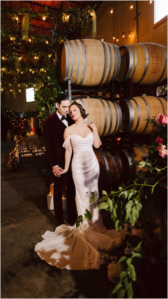 Snohomish Wedding Photographer GSquared Weddings A couple stands in a romantic winery setting. The woman is in a white gown, and the man is in a dark suit. They hold hands near a stack of wooden barrels, surrounded by warm lighting and lush greenery. Serving Seattle, Snohomish and Orlando