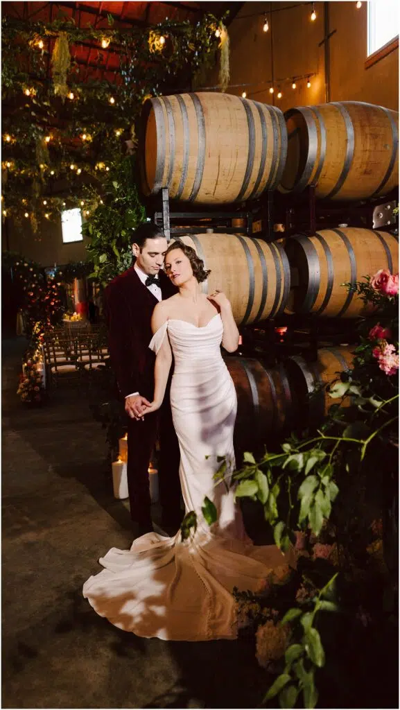 Snohomish Wedding Photographer GSquared Weddings A couple stands in a romantic winery setting. The woman is in a white gown, and the man is in a dark suit. They hold hands near a stack of wooden barrels, surrounded by warm lighting and lush greenery. Serving Seattle, Snohomish and Orlando
