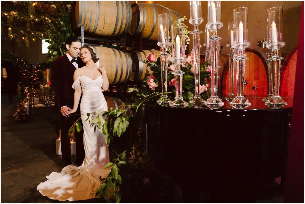 Snohomish Wedding Photographer GSquared Weddings A couple in formal attire stands beside a black grand piano adorned with flowers and lit candles. Behind them are wine barrels and greenery, creating a romantic ambiance. The setting appears to be a winery or cellar. Serving Seattle, Snohomish and Orlando